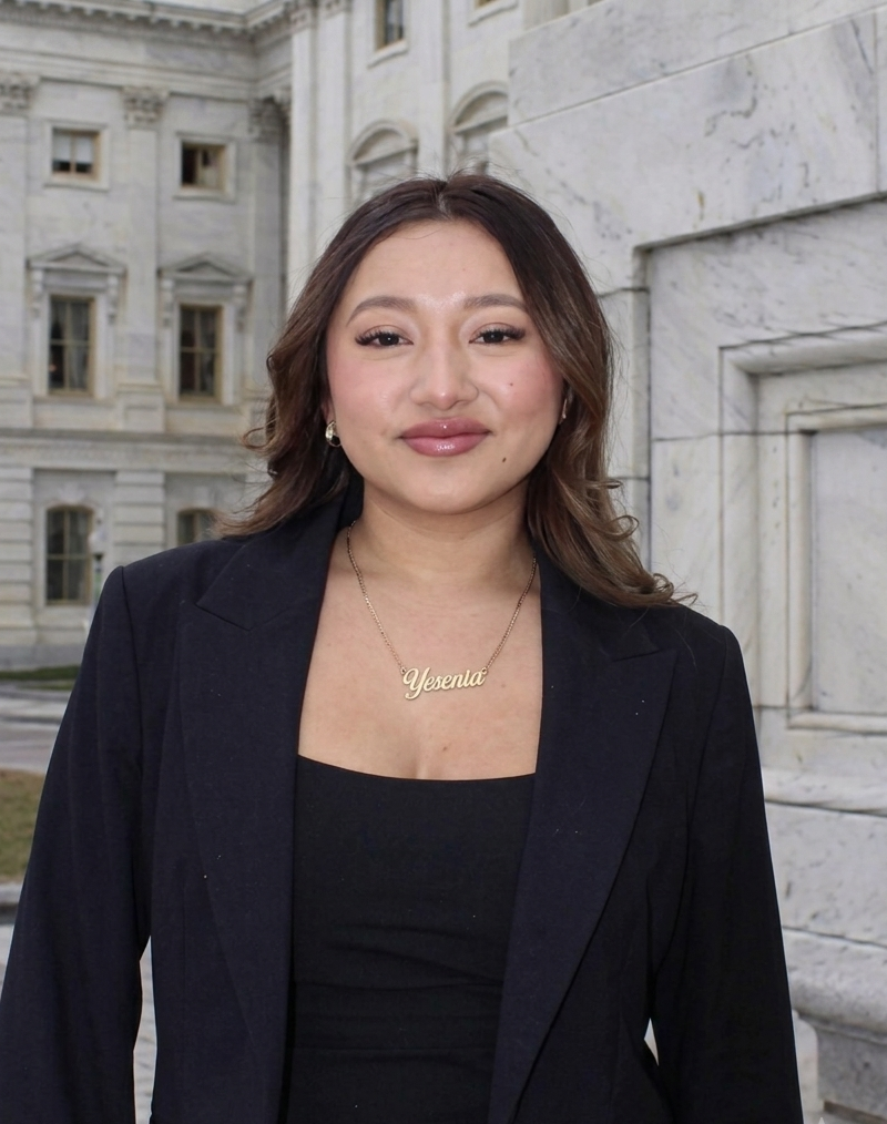 A professional portrait of a smiling woman in a black blazer and necklace, standing outside a white marble government building.